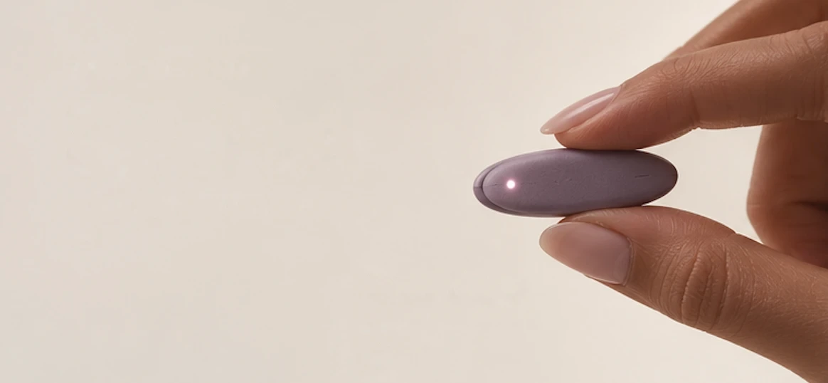 A hand holding Kaṇ̃a, a small plum pebble-shaped wearable by Stri Health